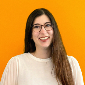 Portrait einer jungen laechelnden Frau mit langen dunklen Haaren und einer Brille auf orangenem Hintergrund.