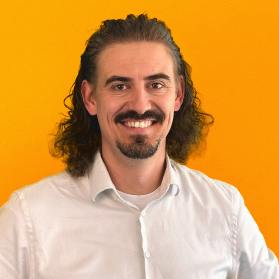 Portrait eines laechelnden Mannes mit langen Haaren und weissem Hemd auf orangenem Hintergrund.