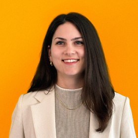 Portrait einer jungen laechelnden Frau mit langen dunklen Haaren und Blazer auf orangenem Hintergrund.