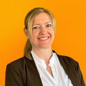 Portrait einer laechelnden Frau mit blonden Haaren und Blazer auf orangenem Hintergrund.