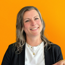 Portrait einer laechelnden Frau mit hellen Haaren und Blazer auf orangenem Hintergrund.